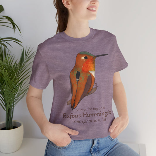 Dark haired woman wearing a purple t-shirt with a Rufous Hummingbird illustration and text, standing indoors next to a potted plant.