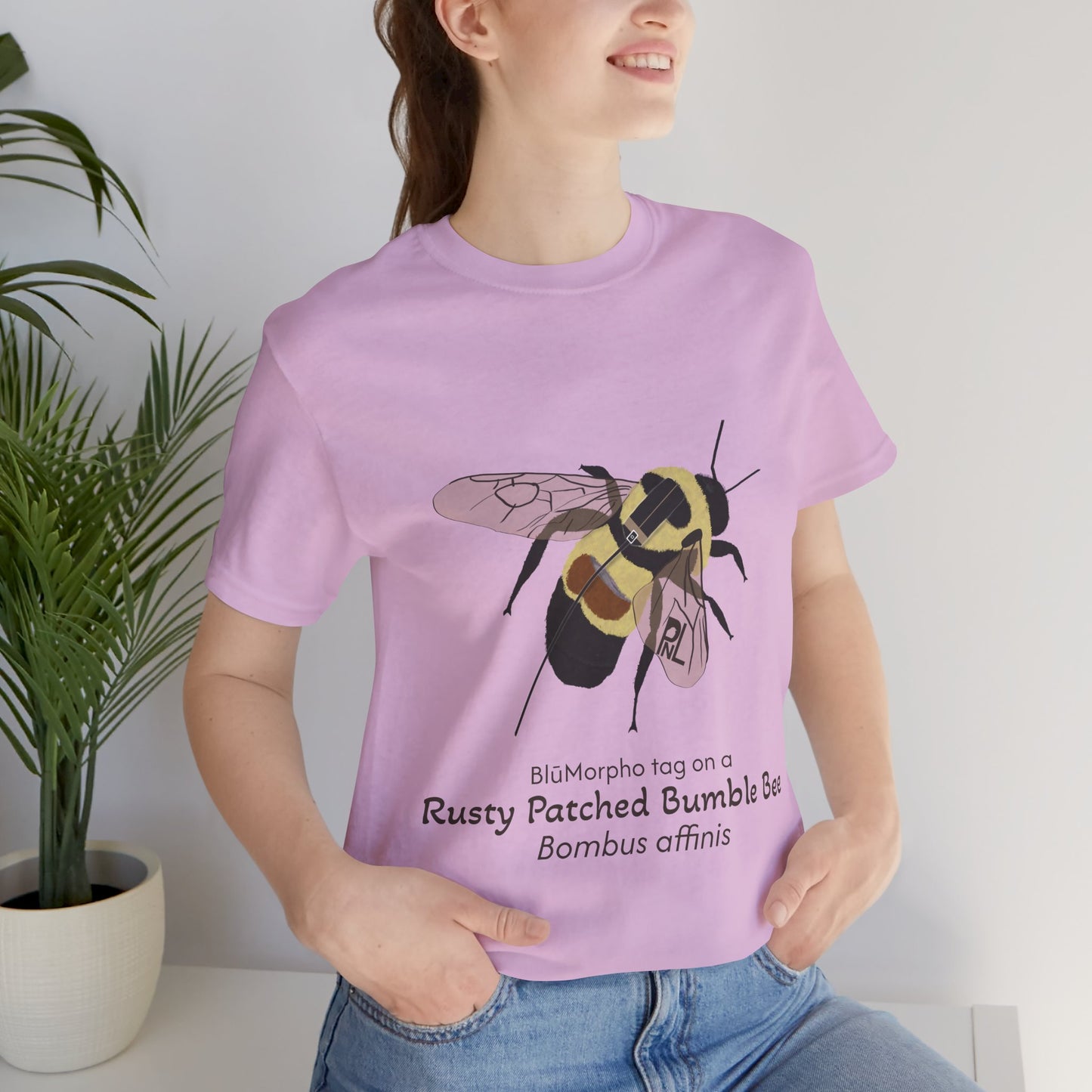 light haired woman wearing a light purple t-shirt with a rusty patched bumble bee illustration and text, standing indoors with a potted plant behind her.