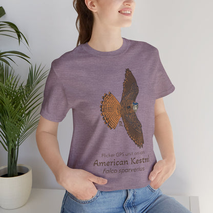 Dark haired woman wearing a purple t-shirt with an American kestrel illustration and text, standing indoors next to a potted plant.