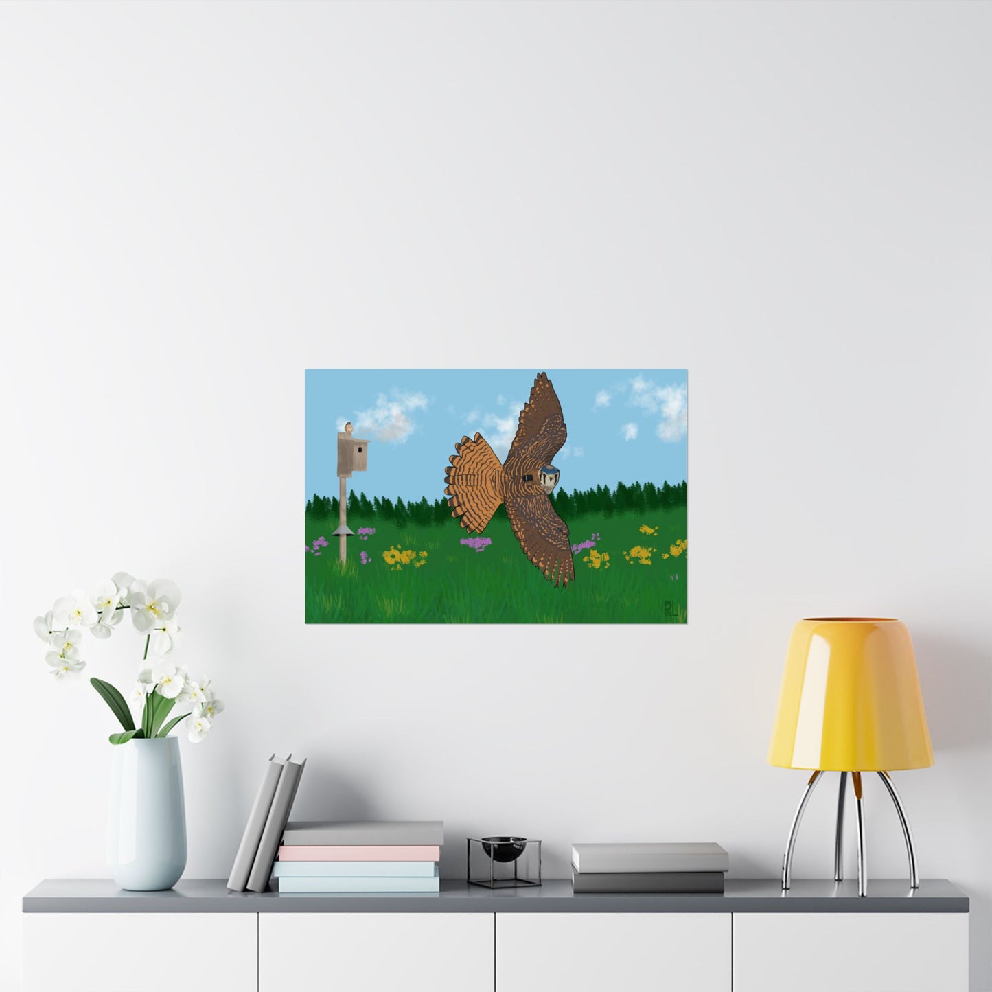 A poster of an American Kestrel flying over a field with flowers and a nest box with a blue sky and forest, on a white wall above a shelf.