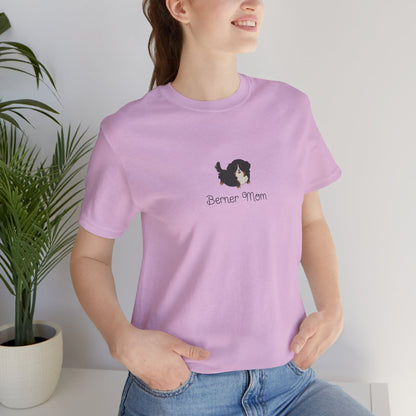 A brown haired woman wearing a purple t-shirt with a kawaii dog illustration and 'Berner Mom' text, standing indoors with a potted plant behind her.