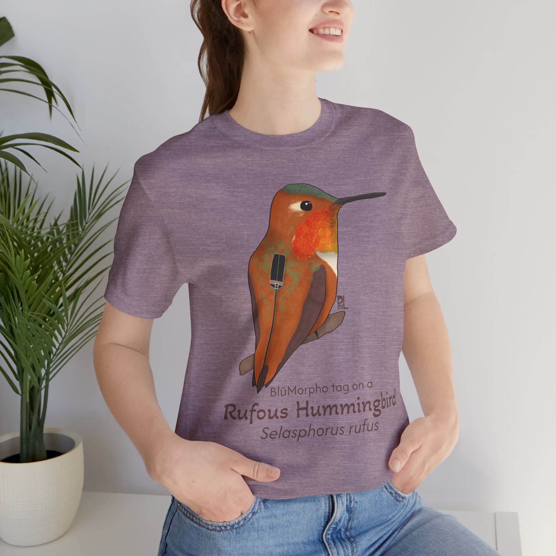 Dark haired woman wearing a purple t-shirt with a Rufous Hummingbird illustration and text, standing indoors next to a potted plant.