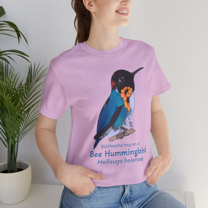 dark haired woman wearing a light purple t-shirt with a colorful bee hummingbird illustration and text, standing indoors next to a potted plant