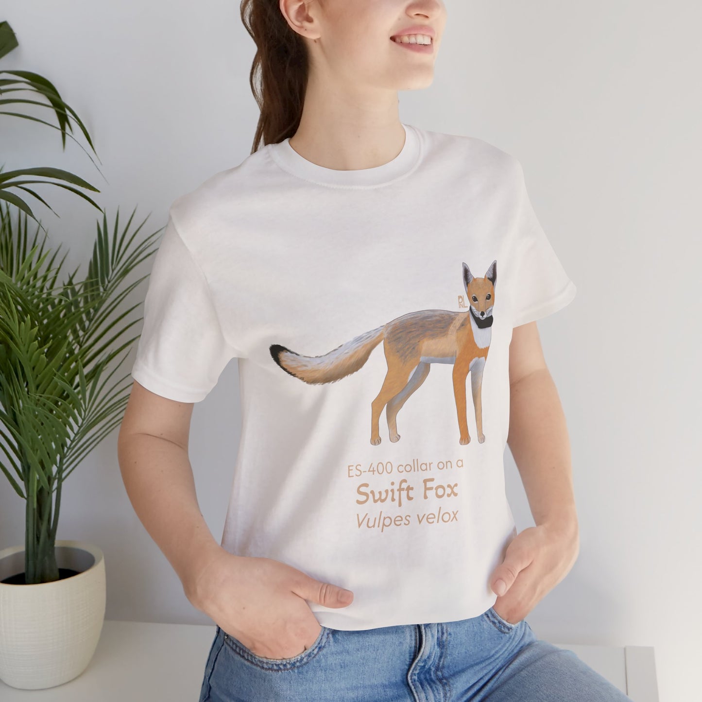 dark haired woman wearing a white t-shirt with a swift fox illustration and text, standing indoors next to a potted plant.