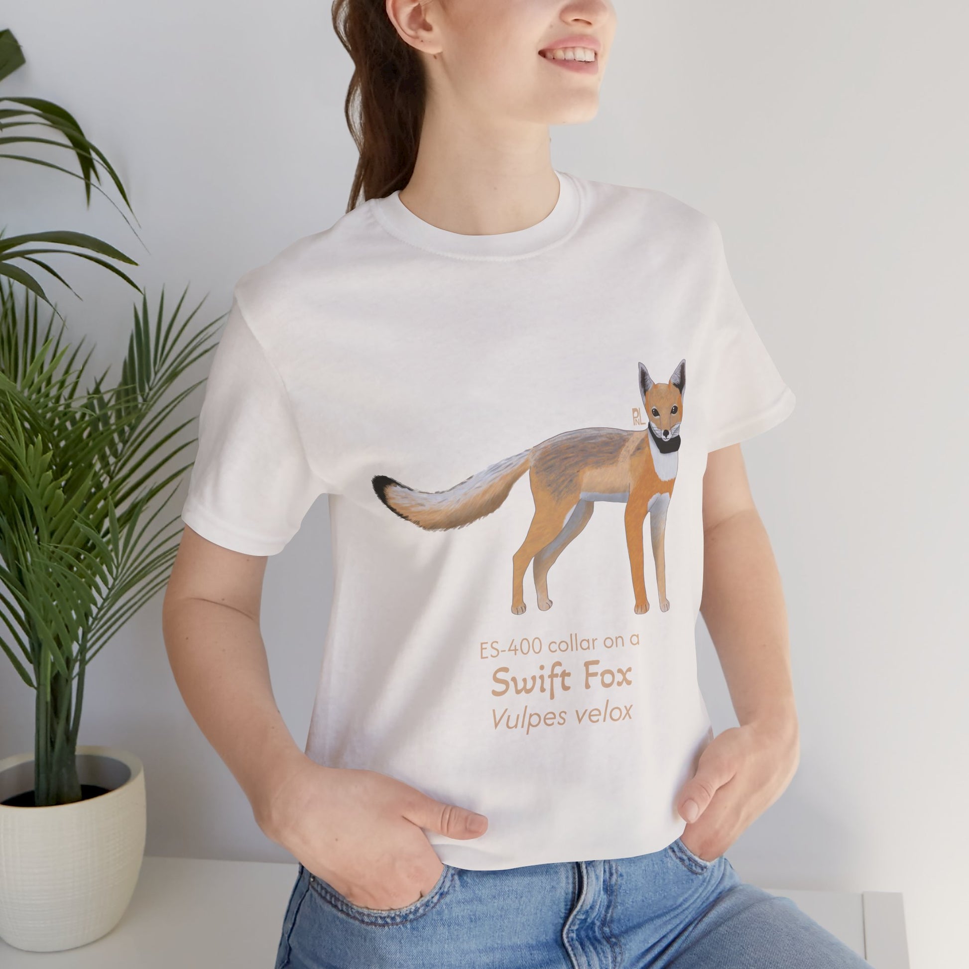 dark haired woman wearing a white t-shirt with a swift fox illustration and text, standing indoors next to a potted plant.
