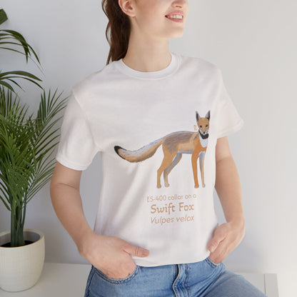 dark haired woman wearing a white t-shirt with a swift fox illustration and text, standing indoors next to a potted plant.