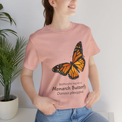 Dark haired woman wearing a light orange t-shirt with a monarch butterfly illustration and text, standing indoors next to a potted plant